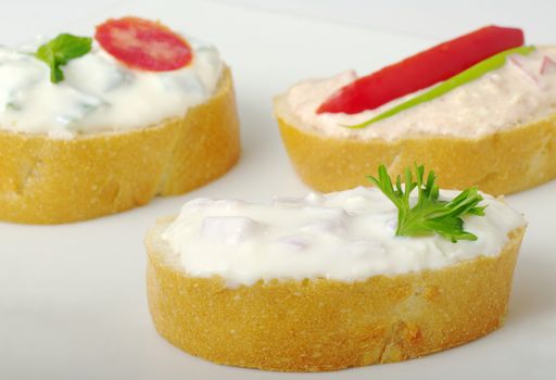 Baguette bread with cream cheese spread decorated with tomato, red pepper, parsley and green onion on white plate (selective focus)
