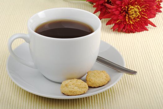 Black tea in cup with saucer, teaspoon, biscuits and flower in background on tablemat (Selective focus)