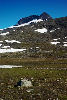 Mountain on the Nordkalottleden in Scandinavia with small snowfields, a tiny lake and some trail markers with blue sky 