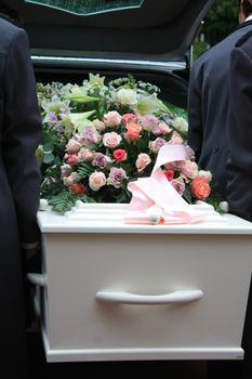 A white coffin, covered with flowers near a hearse, people taking out the coffin