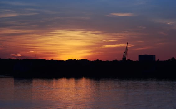The silhouette of a crane and an industrial building on the shore at sunset 