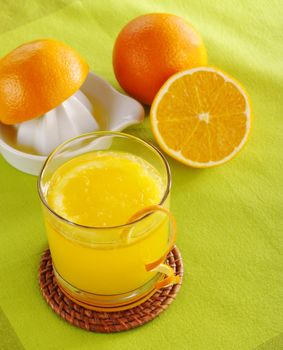 Fresh orange juice with orange slice in glass and squeezer with oranges in background on green table mat (Selective Focus)