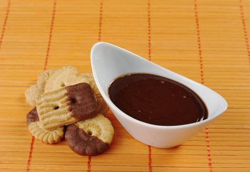 Butter cookies dipped in milk chocolate icing with chocolate dip in white bowl on orange-colored table mat (Selective Focus, Focus on front rim of bowl and part of the surface of the icing as well as the front cookie)  