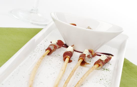 Thin ham slices wrapped around thin bread sticks and served on a long plate on a green place mat with a wine glass in the background (Selective Focus) 