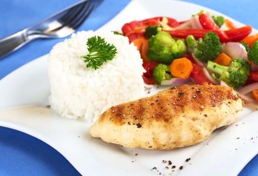 Fried chicken breast with pepper on top, vegetables (carrot, broccoli, onion, red bell pepper) and rice garnished with parsley (Selective Focus, Focus on the front of the meat)