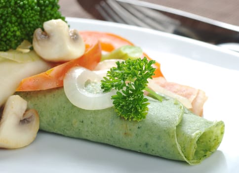Rolled thin green colored pancake (colored with spinach in dough) with vegetables on top with fork in the background (Selective Focus, Focus on the front side of the pancake and the parsley) 