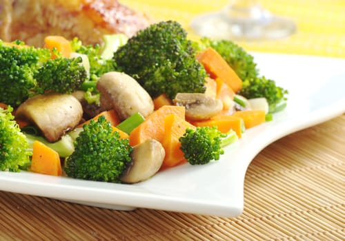 Fried vegetables (broccoli, mushroom, carrot, shallot) on white plate with chicken meat and wine glass in the background (Selective Focus, Focus on the vegetables in the front) 