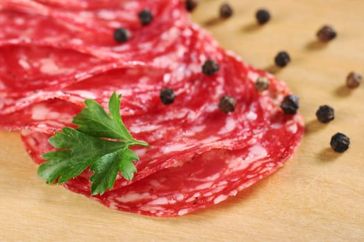 Salami slices with parsley leaf on top and black pepper corns on wooden board (Selective Focus, Focus on the front of the first two slices and the front of the leaf)