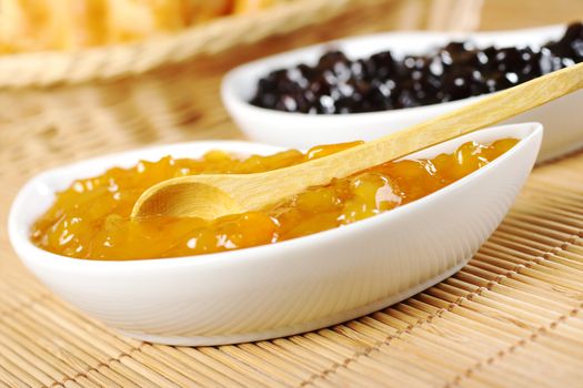 Peach jam with wooden spoon in small white bowl with red jam and bread basket in the background (Selective Focus, Focus on the spoon) 