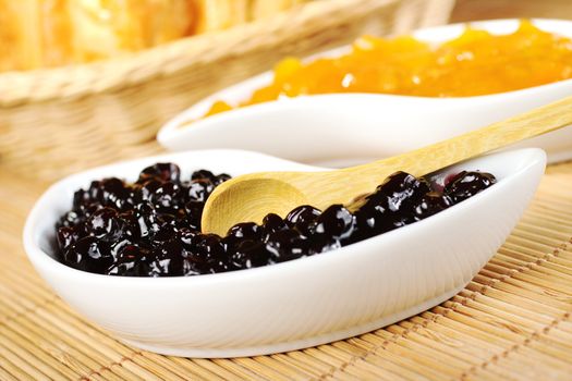 Elder berry jam with wooden spoon in small white bowl with peach jam and bread basket in the background (Selective Focus, Focus on the spoon) 