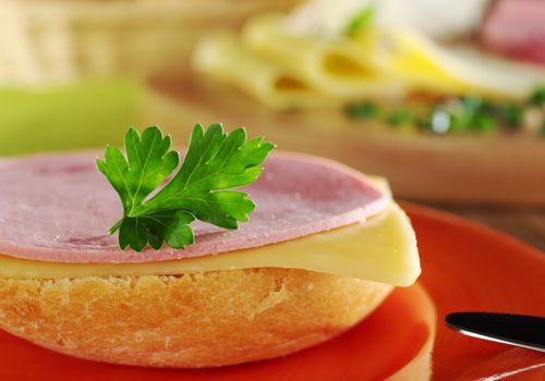 Open sandwich with a cold meat slice and cheese slice with parsley on top on orange plate with knife, and ingredients in the background (Selective Focus, Focus on the front of the bun, the slices and the parsley)
