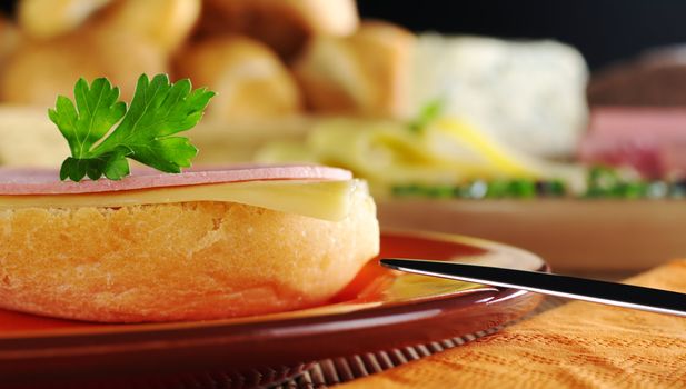 Open sandwich with a cold meat slice and cheese slice with parsley on top on orange plate with knife, and ingredients in the background (Selective Focus, Focus on the front of the bun, the slices and the parsley)