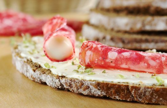 Salami slice and cream cheese with dried herbs on a slice of brown bread on wooden board and ingredients in the background (Selective Focus, Focus on the salami in the front)