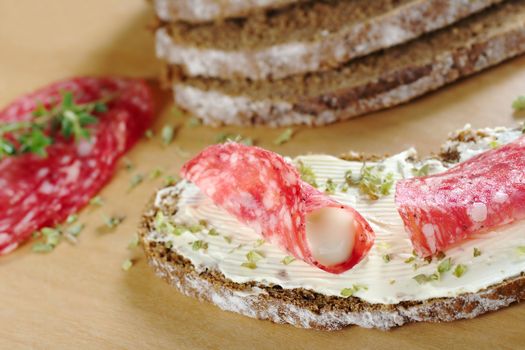 Rolled salami slice filled with cream cheese with dried herbs on a slice of brown bread on wooden board and ingredients in the background (Selective Focus, Focus on the front of the salami roll and filling)