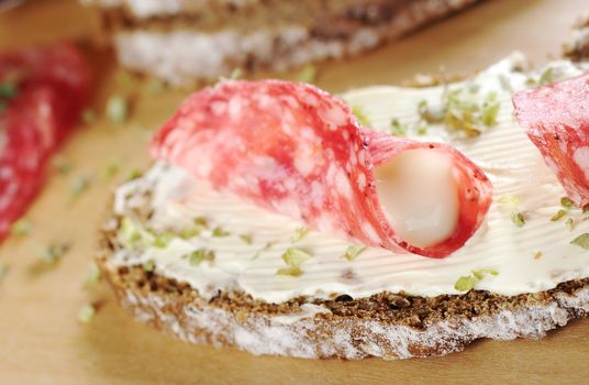 Rolled salami slice filled with cream cheese with dried herbs on a slice of brown bread on wooden board and ingredients in the background (Selective Focus, Focus on the front of the salami roll and filling)