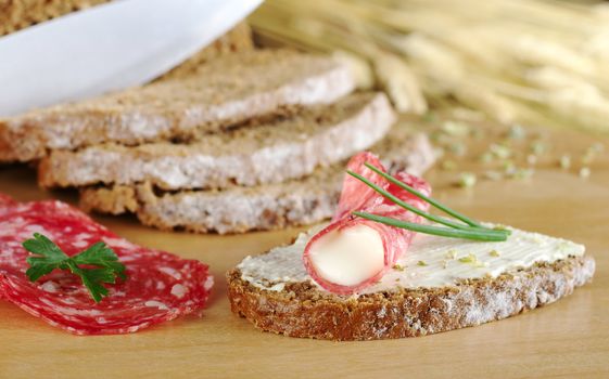 Rolled salami slice and cream cheese with chives on half a slice of brown bread on wooden board and ingredients (Selective Focus, Focus on the roll and bread slice in the front and the front of salami slices on the board)