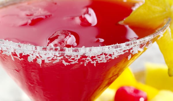 Red cocktail with ice cubes and grated coconut and carambola on the glass rim and fruits in the background (Selective Focus, Focus is on the front rim of the glass)