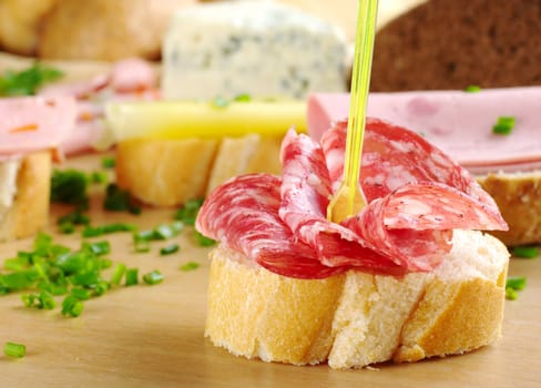 Salami slices on baguette with a plastic skewer and other sandwiches in the background on a wooden board (Selective Focus, Focus on the plastic skewer)