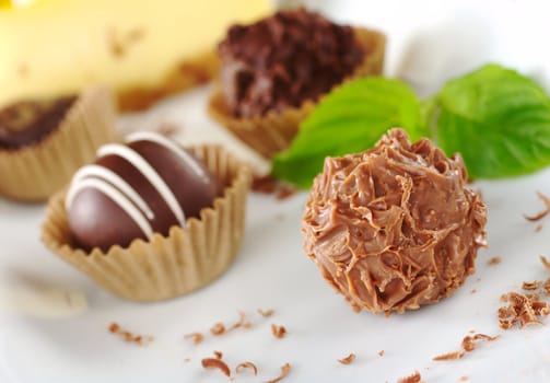 Chocolate truffles with mint leaf, fine chocolate shavings and a yellow cake in the background (Very Shallow Depth of Field, Focus on the front of the first praline) 