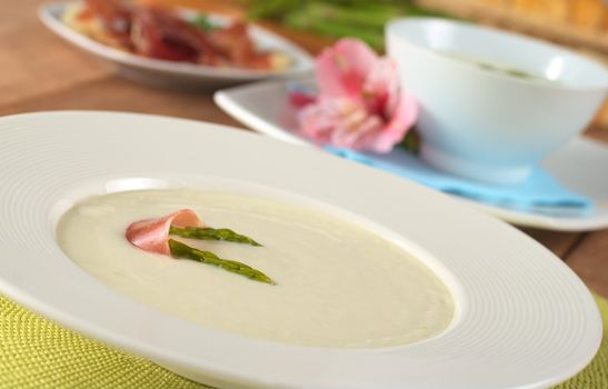 Cream of asparagus garnished with green asparagus heads and a slice of ham on top in a white soup plate (Selective Focus, Focus on the asparagus head in front and part of the ham slice on the soup)