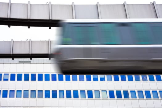 Modern public transportation system skytrain hanging from elevated guideway passes by in front of tall office building facade (with motion blur).