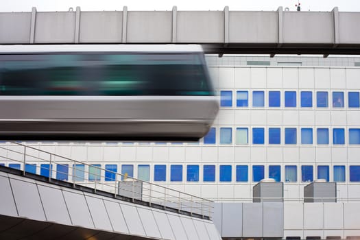 Modern public transportation system skytrain hanging from elevated guideway passes by in front of tall office building facade (with motion blur).