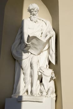 Sculpture of old shepherd with huge book in hands and animal near legs looking at him.