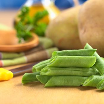 Raw cut green bush beans on wooden board with a kitchen knife, potatoes, parsley and oil in the back (Selective Focus, Focus on the three beans lying on each other) 