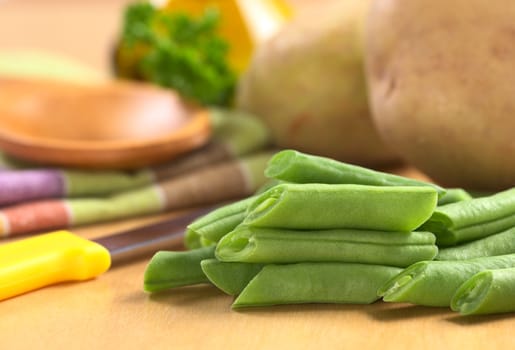 Raw cut green bush beans on wooden board with a kitchen knife, potatoes, parsley and oil in the back (Selective Focus, Focus on the three beans lying on each other) 