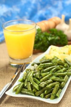 Cooked green beans with onion and parsley with cooked potato, orange juice and ingredients in the back (Selective Focus, Focus on the front of the beans)