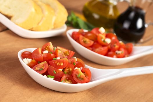 Cherry tomato wedges with cheese and chives prepared as salad served in ceramic spoons with baguette slices, oil and balsamic vinegar in the back (Selective Focus, Focus on the salad in the front)