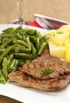 Beef meat slices with fresh rosemary on top, served with green beans and cooked potatoes (Selective Focus, Focus on the front of the upper meat slice and the front of the rosemary) 