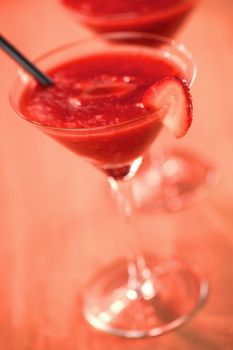 Frozen Strawberry Daiquiri Made of rum, strawberries, ice, sugar and lemon juice served in a cocktail glass with a strawberry slice on the rim and a black straw photographed with a red lighting filter on wood (Very Shallow Depth of Field, Focus on the front of the glass rim and the front top of the strawberry slice on the rim)