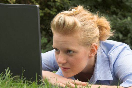 Closeup portrait of a pretty blonde laying on the grass and looking at laptop screen