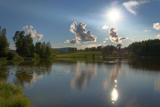 A still lake in the late summer with the Sun, clouds and a school on a hill side. The image is HDR tonemapped from 3 exposures with detail enhancement.

