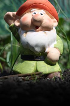 A small decorative handmade garden gnome with an orange hat a white beard and a green tunic. Set against a backdrop of a vegetable patch.