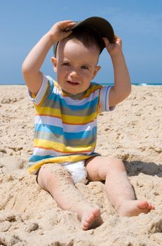 One year old boy sitting on sand with cap in hands