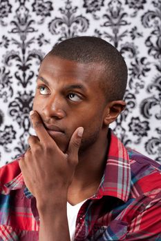 An African American man with his hand on his chin thinking deeply about something in front of a damask style background. Shallow depth of field.