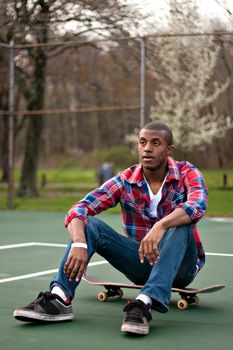 A young man hanging out in the tennis courts sitting on his skateboard.