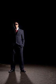  An African American man dressed in a dark colored suit and sunglasses standing in front of a dark black background.