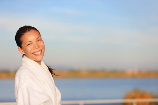 Spa resort woman smiling wearing bathrobe outdoors with water view on lake. Beautiful smiling happy mixed race Chinese Asian / white Caucasian female model outside.