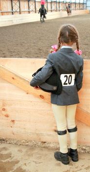 a young female equestrian with riding helmet under her arm, patiently waits to be called to compete in the show ring