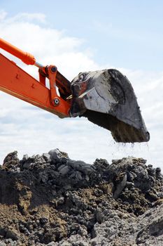 Bucket of orange mechanical digger dropping a scoop of clay soil on heap of dirt during major rural construction.