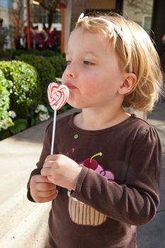 Cute little European toddler girl having fun eating heart shaped lollypop