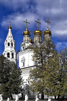 White Stone Holy Trinity Cathedral with gold baths and crosses against the blue sky, white clouds and yellow-green trees (Verhoturie city of Sverdlovsk Region)