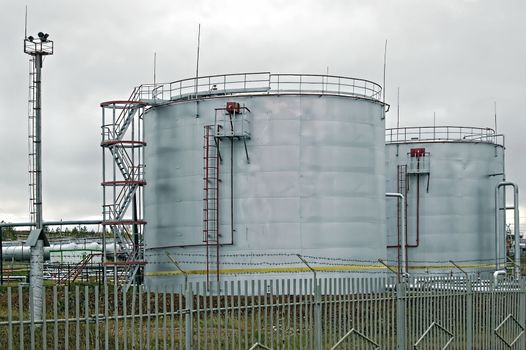 Silvery-gray oil storage tanks with a red ladder and railings, communications tower, pipe, fence in the background of yellow-green grass and gray skies