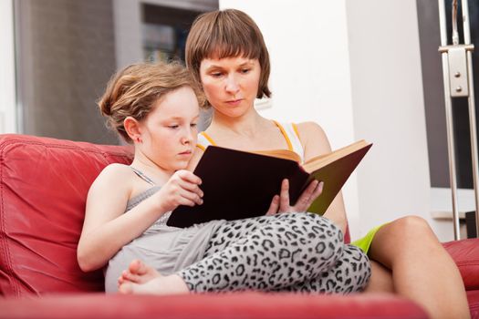 Mother and teenager daughter sitting on sofa together and reading an old book