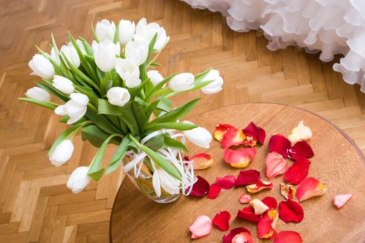 Vase with white tulips on a wooden little table and petals of a red tulip beside