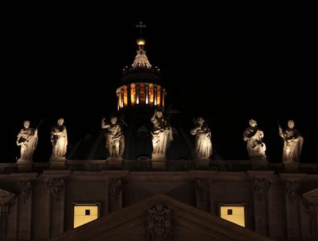The top of St. Peter's Basilica, in Vatican city at night, featuring the statues and the top of it's dome.
