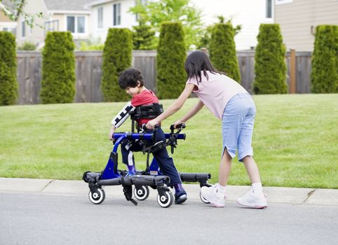 Big sister helping younger disabled brother to walk in his walker outside